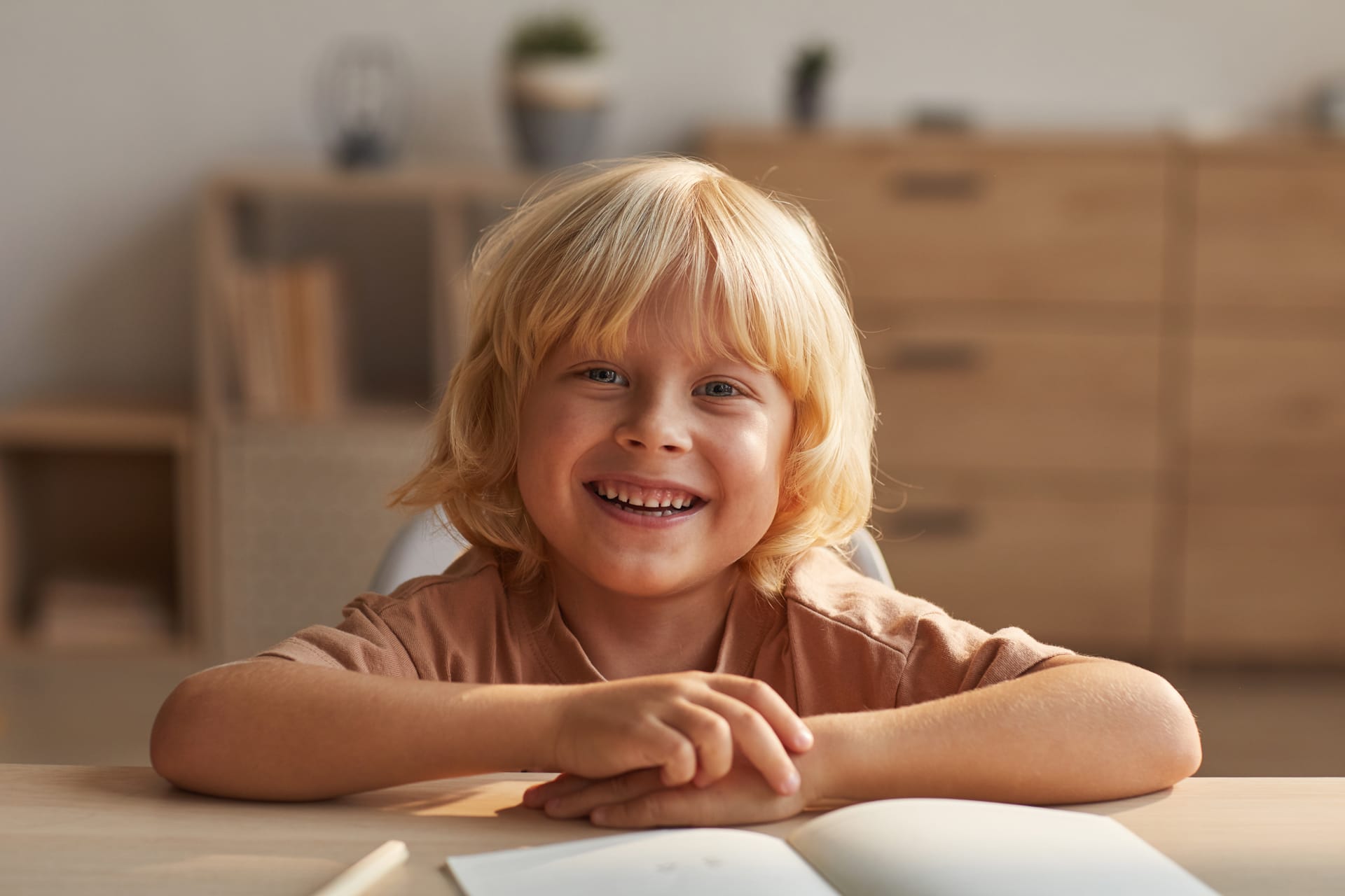 Boy with blond hair smiling while sitting table with copybooks