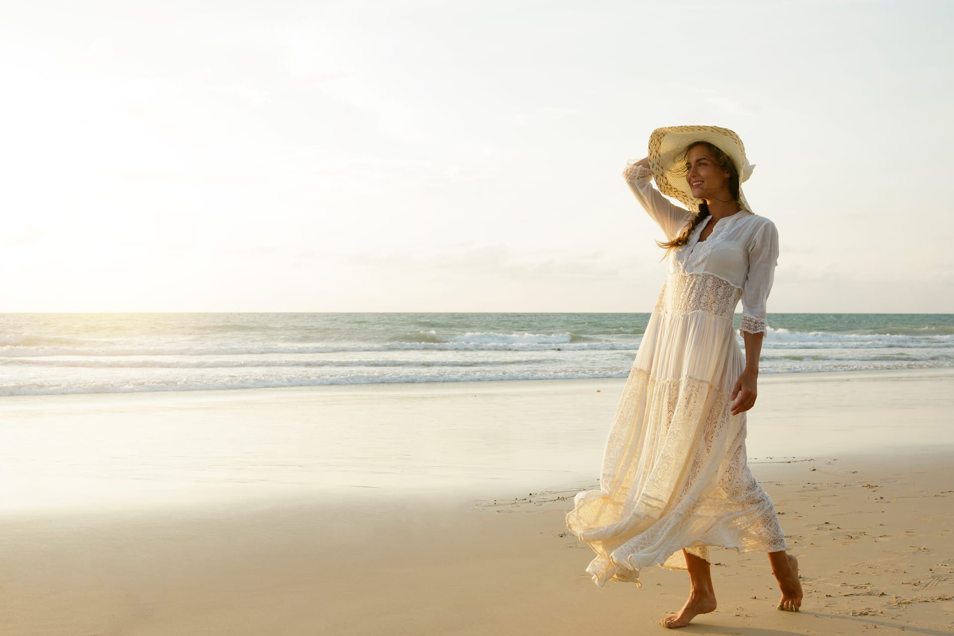 Woman wearing beautiful white dress is walking beach during sunset