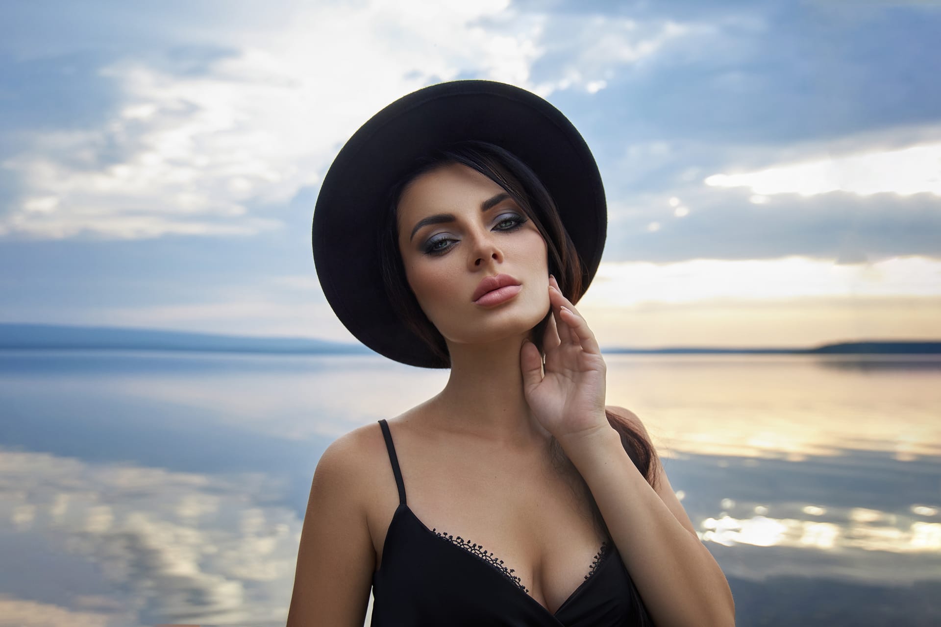 Brunette beauty woman black hat black dress poses near lake against blue sky