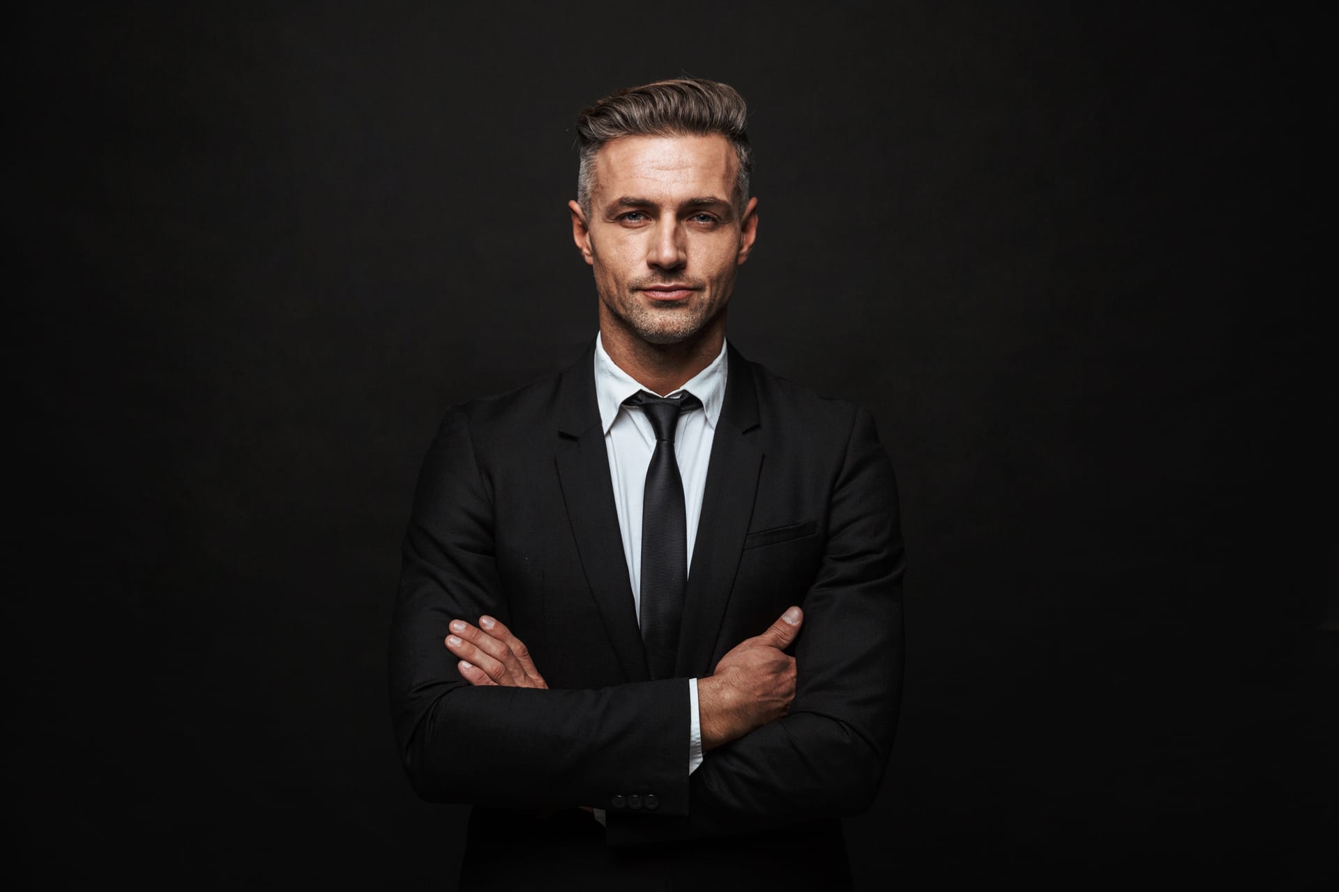 Handsome man wearing suit standing isolated black wall arms folded