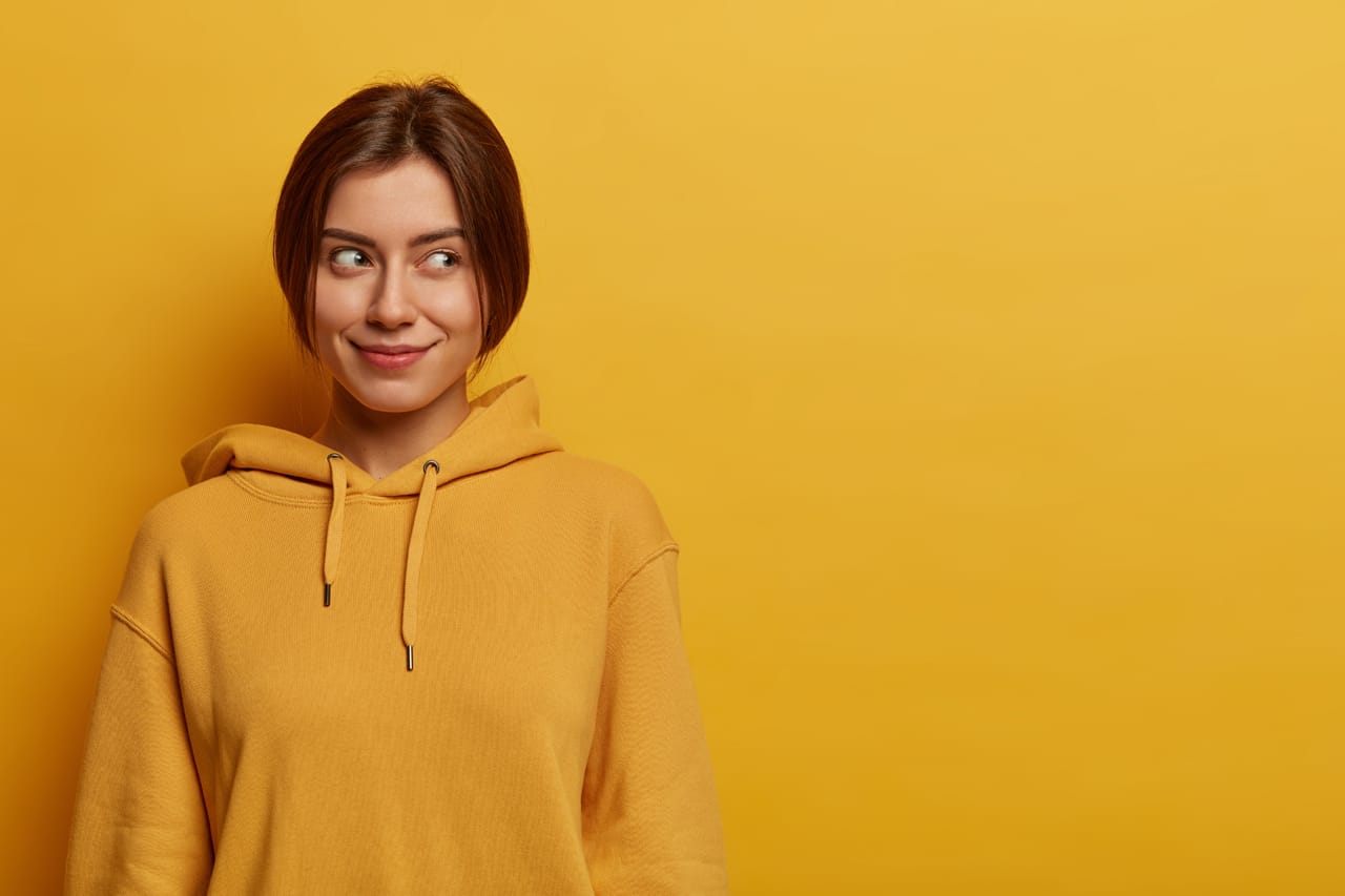 Related image portrait dreamy satisfied young european woman looks aside blank space has pleasant look wears hoodie poses against yellow wall